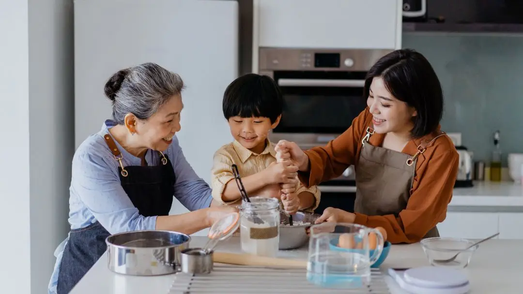 home-insurance-family-cooking-singapore Grandparent, child, and adult cooking together in a modern kitchen, representing family bonding and the importance of protecting home and lifestyle in Singapore.